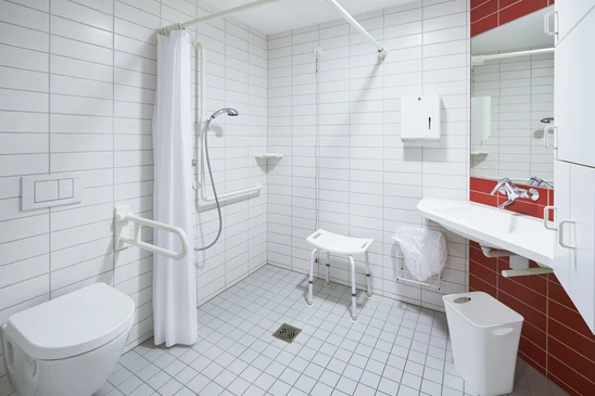 Salle de bain adaptée PMR avec douche à l'italienne équipée de barres d'appui murales blanches, siège de douche rabattable et tabouret blanc réglable
