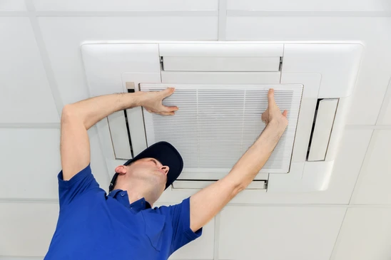 Un technicien en uniforme bleu et casquette noire installe ou remplace un filtre à air dans un système de ventilation plafond intérieur blanc